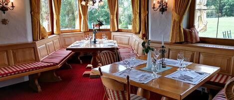 A dining area with a wooden table set for four, benches along the walls, and large windows with curtains.