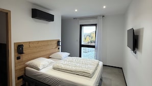 Ein modernes Schlafzimmer mit einem Holz-Kopfteil, einem großen Bett mit weißer Bettwäsche, einem an der Wand montierten Fernseher und einem Fenster mit Blick auf Bäume.