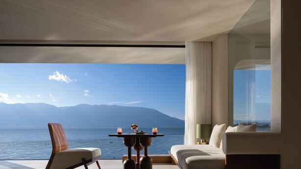 Habitación moderna con vistas a un lago y montañas, un sofá, una silla y una mesita con bebidas.