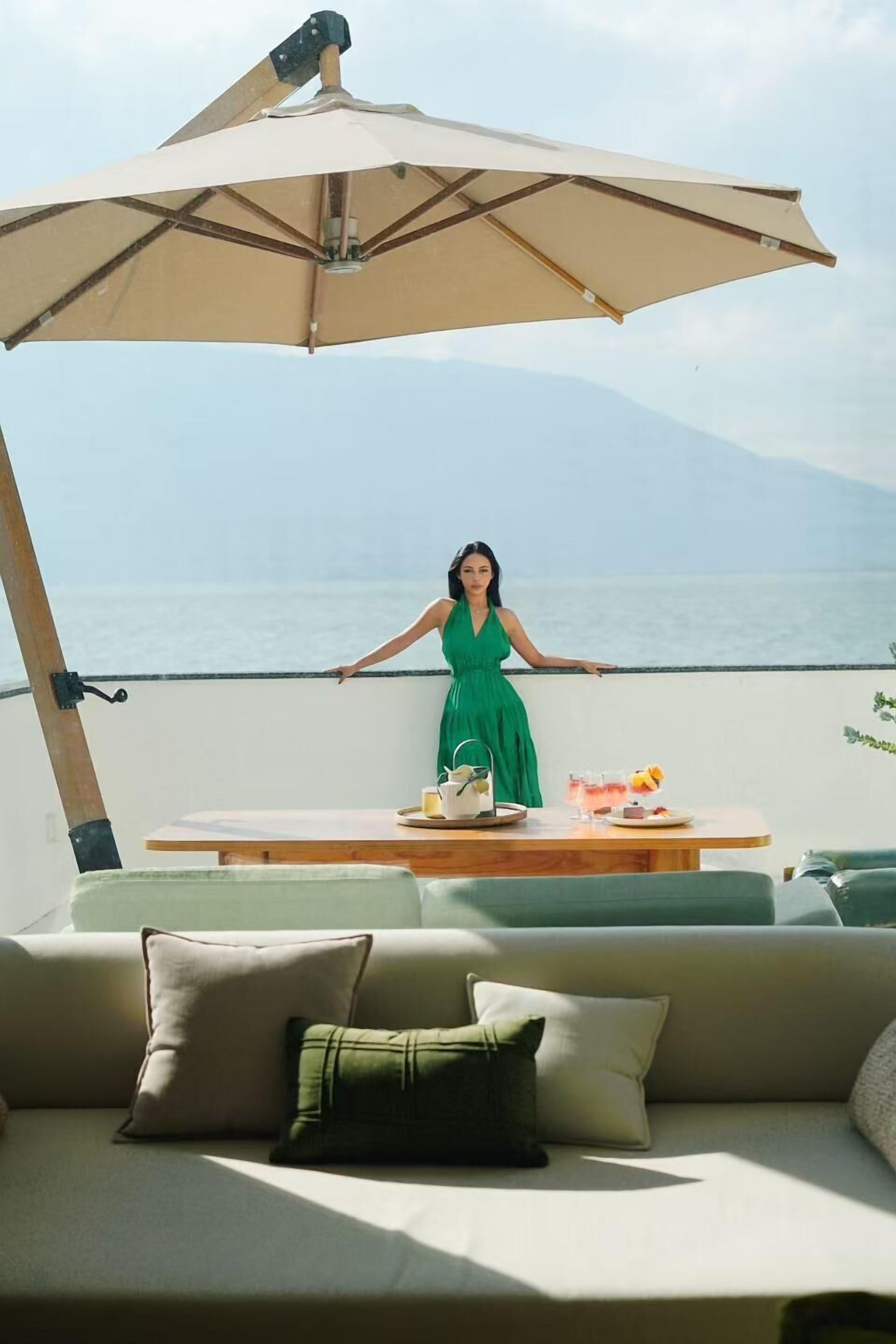 Una persona con un vestido verde está de pie junto a una mesa con bebida y comida, con vista a una playa y montañas al fondo.