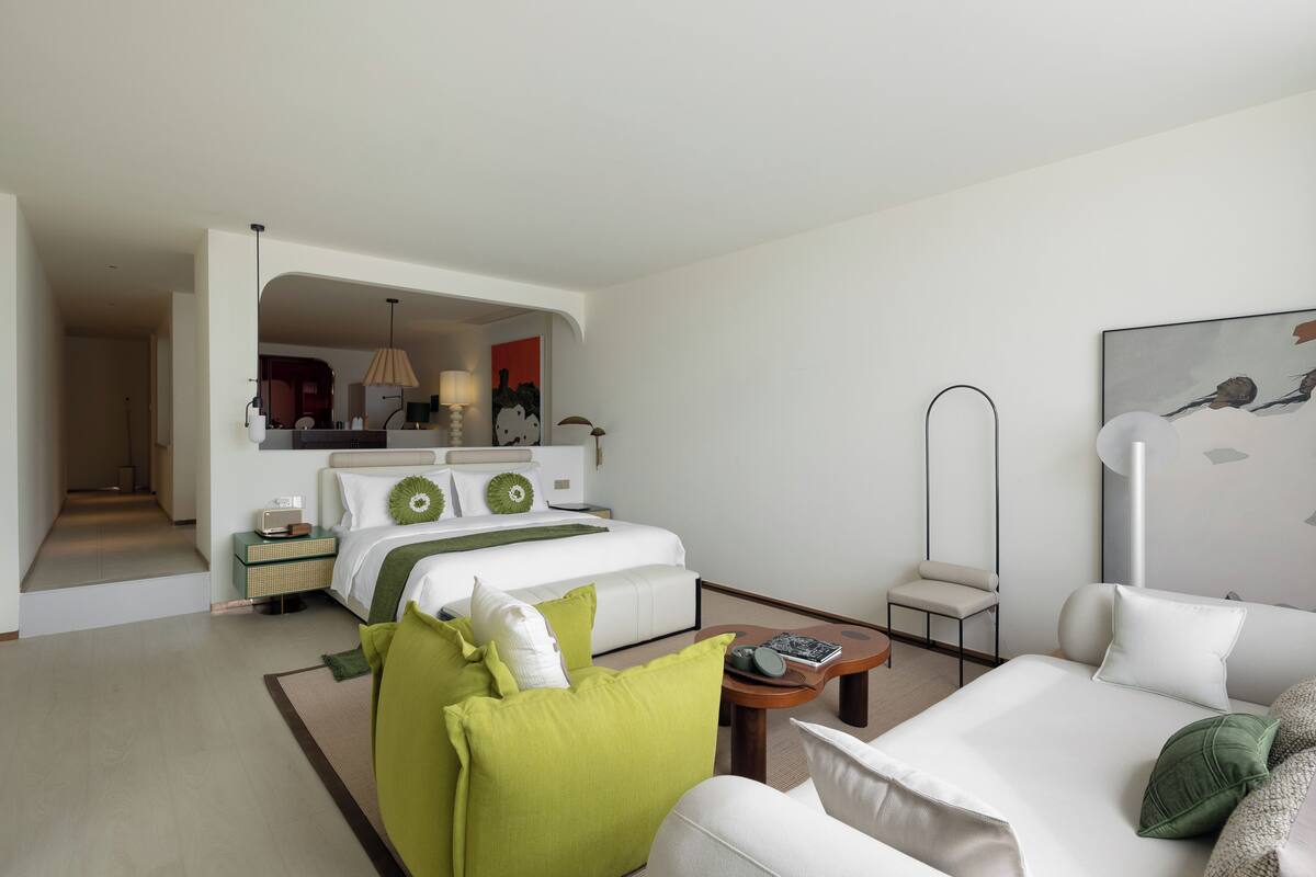 Ein Hotelzimmer mit einem großen Bett, einer Couch, einem kleinen Tisch und einem Sessel. An der Wand hängt ein Spiegel und über dem Bett ein Gemälde.