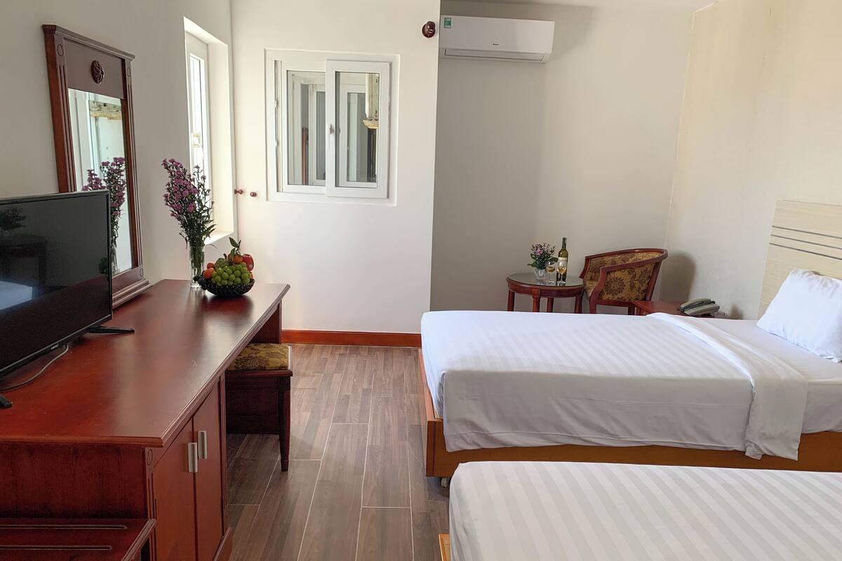 A hotel room with two beds, a TV, a desk with a computer, a chair, a small table with a vase of flowers, and an air conditioning unit.