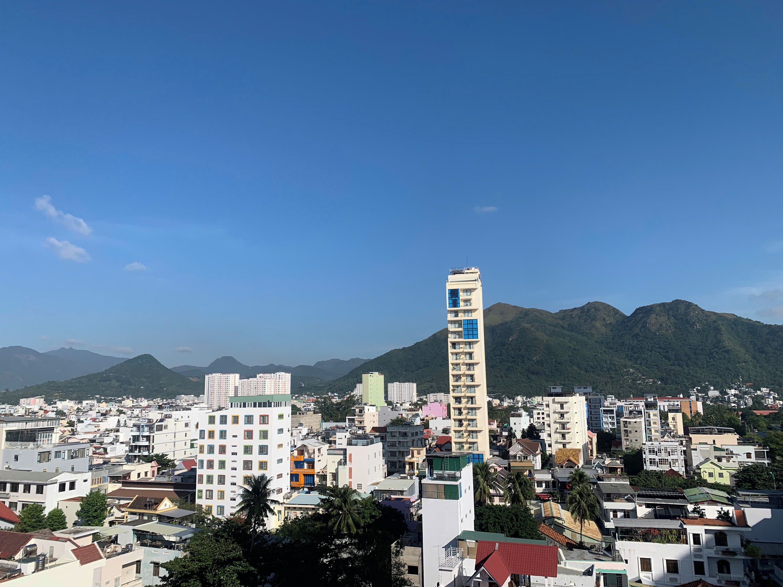 Un paisaje urbano con un edificio alto y destacado que posee múltiples balcones, rodeado de edificios residenciales y montañas al fondo.