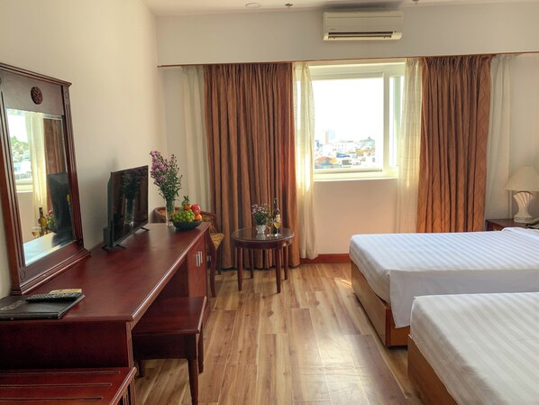 Een hotelkamer met een houten bureau, twee bedden, een kleine tafel met een bloemenvaas, een raam met gordijnen en uitzicht op de stad.