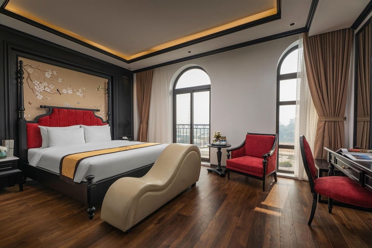 Habitación de hotel con una cama grande, un sillón rojo, un escritorio con una silla y vistas a la ciudad a través de amplios ventanales.