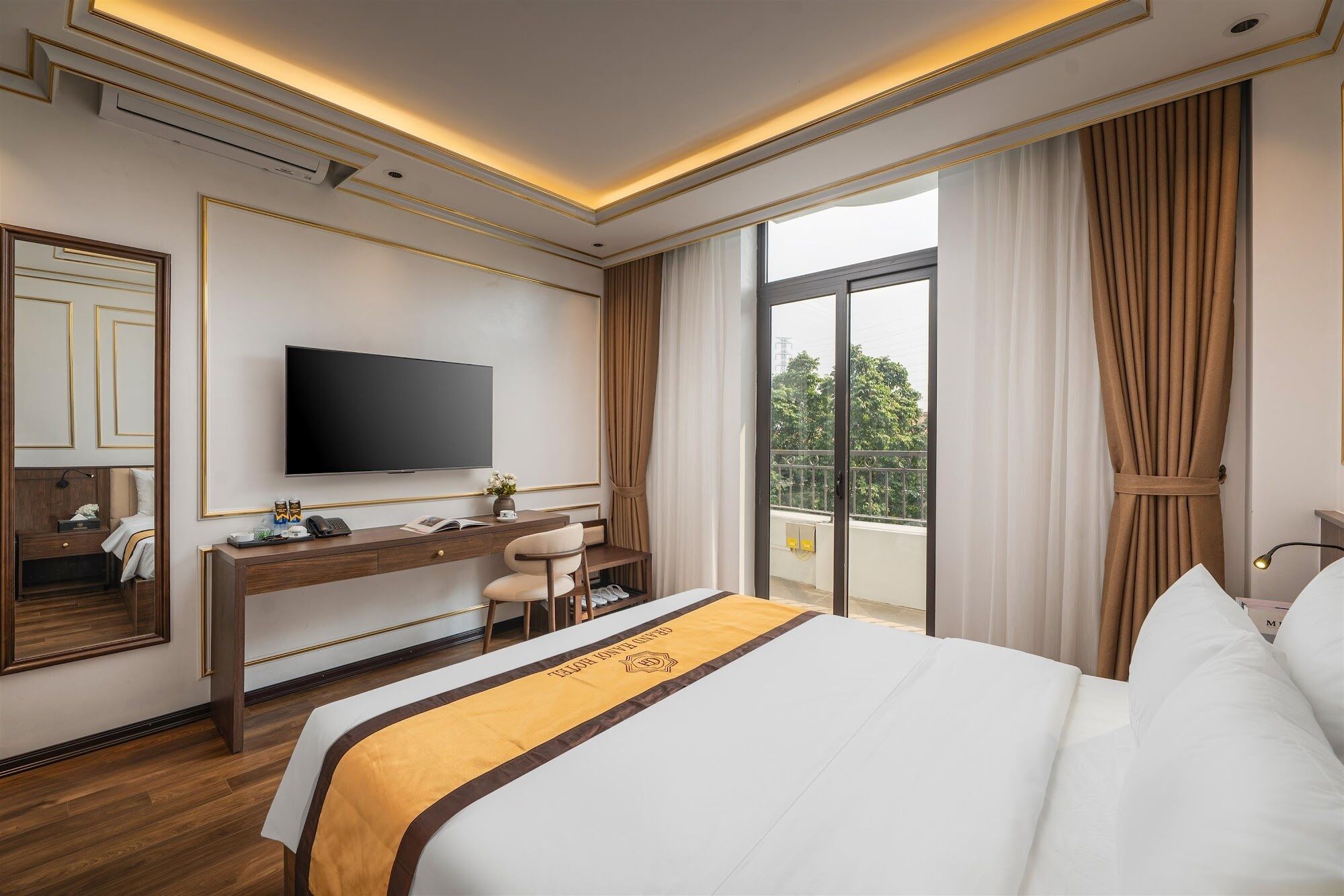 A hotel room with a large bed, a desk with a flat-screen TV, a chair, and a balcony with curtains.