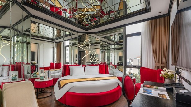Une chambre d’hôtel moderne avec un lit rond, une décoration rouge et blanche, un bureau avec un magazine et de grandes fenêtres donnant sur la ville.