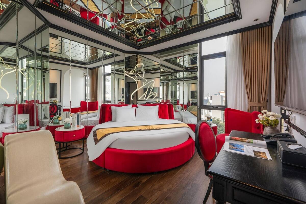 Una habitación de hotel moderna con una cama redonda, decoración en rojo y blanco, un escritorio con una revista y ventanas amplias que ofrecen vistas a la ciudad.