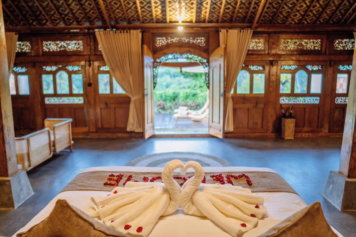 A room with a bed, a wooden ceiling, and a view of greenery outside.