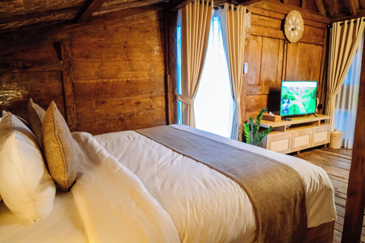 A wooden cabin room with a bed, curtains, a television, and a wooden cabinet.
