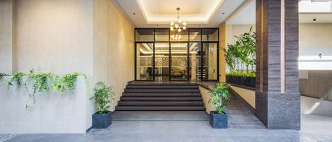 A modern hotel entrance with a staircase leading to a reception area, featuring a geometric ceiling design and potted plants.