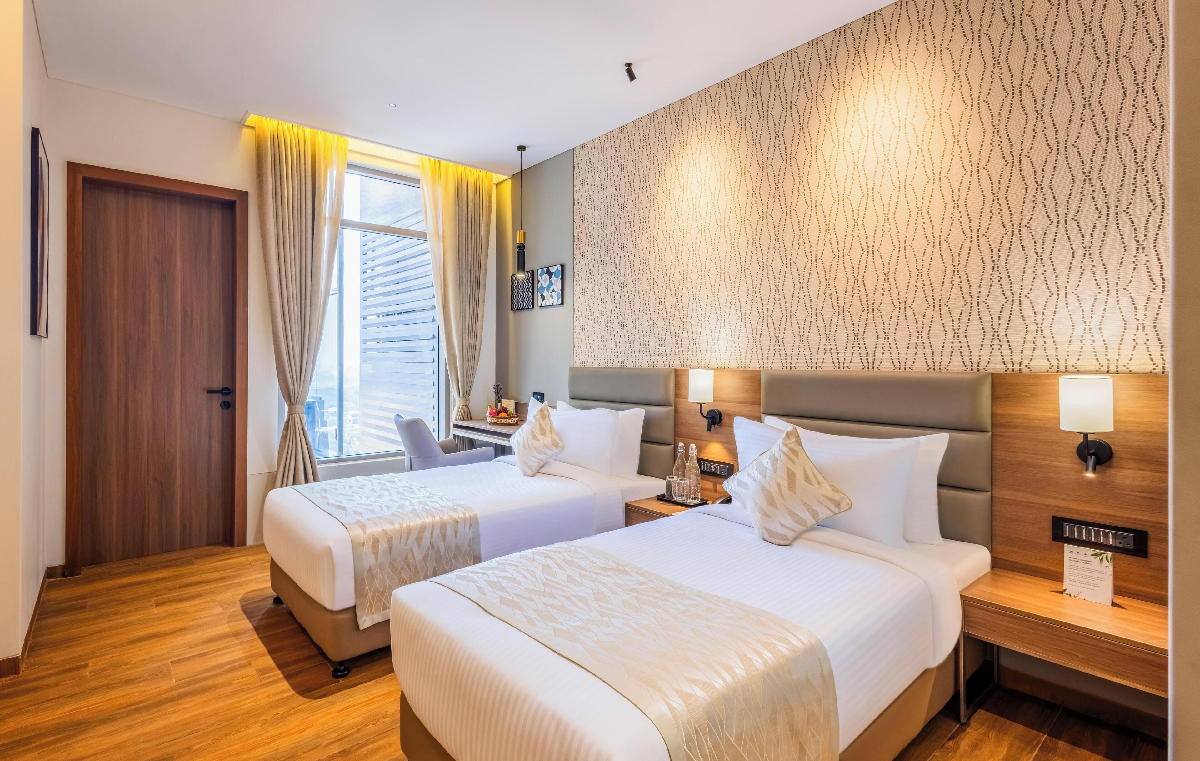 A hotel room with two beds, a wooden floor, a patterned wall, and a window with curtains.