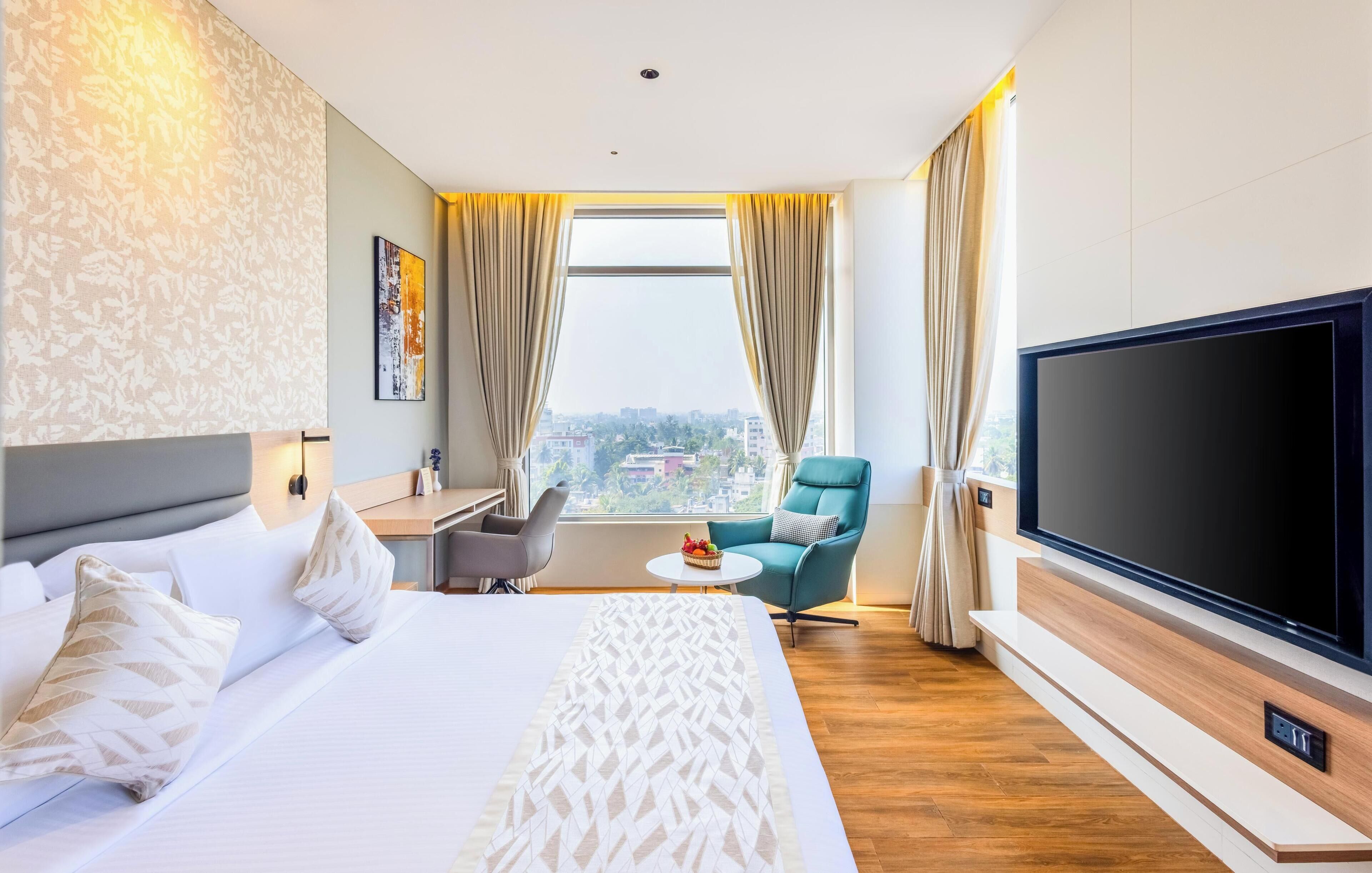 A hotel room with a large bed, a TV, a desk, a chair, and a window with a city view.