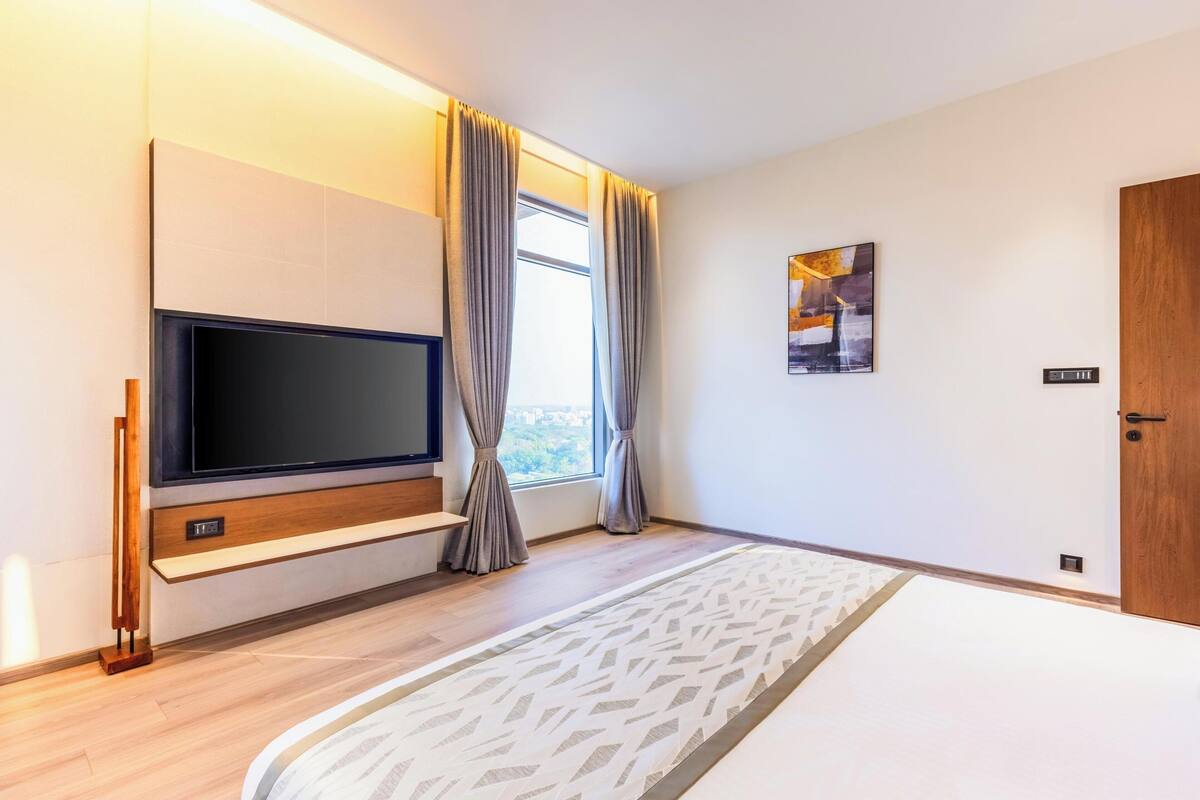 A modern hotel room with a large flat-screen TV, a wooden TV stand, a window with curtains, a patterned rug, and a door.