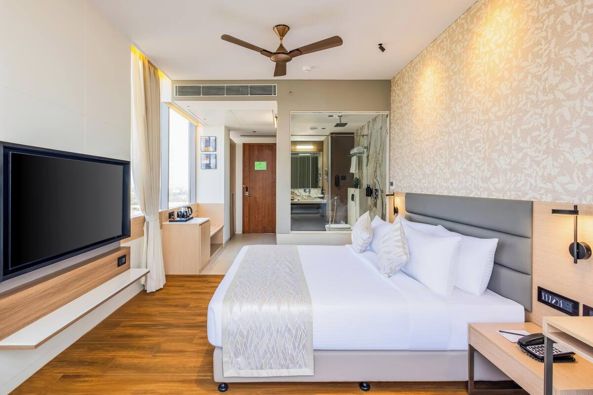 A modern hotel room with a large flat-screen TV, a bed with white linens, a ceiling fan, and a bathroom visible through an open door.