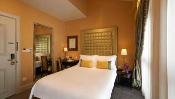 Habitación de hotel con una cama grande, cabecera, mesita de noche con lámpara, espejo y ventana con cortinas.