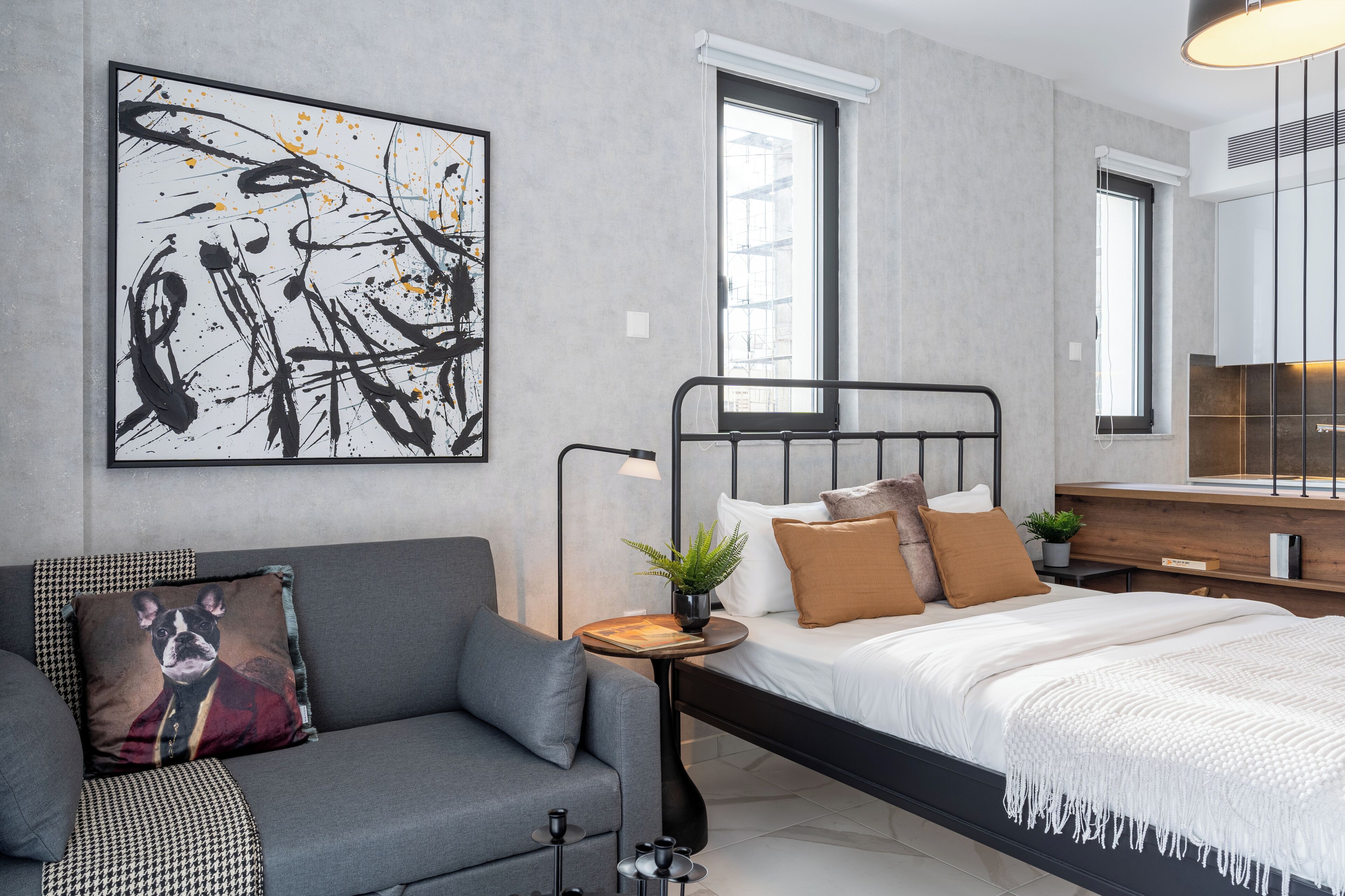 A modern bedroom with a bed, a grey sofa, a wall-mounted artwork, and a bedside table with a plant.