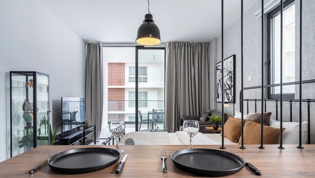 Un espace repas moderne avec une table en bois dressée pour deux personnes, un luminaire suspendu noir et une vue sur un balcon donnant sur le paysage urbain.