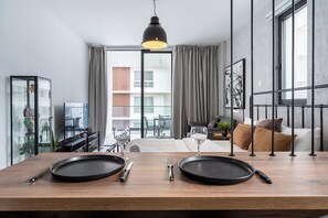 A modern dining area with a wooden table set for two, a black pendant light, and a view of a balcony with a cityscape. - Dusk - Studio by Plus 49 in Mackenzie (Larnaca)