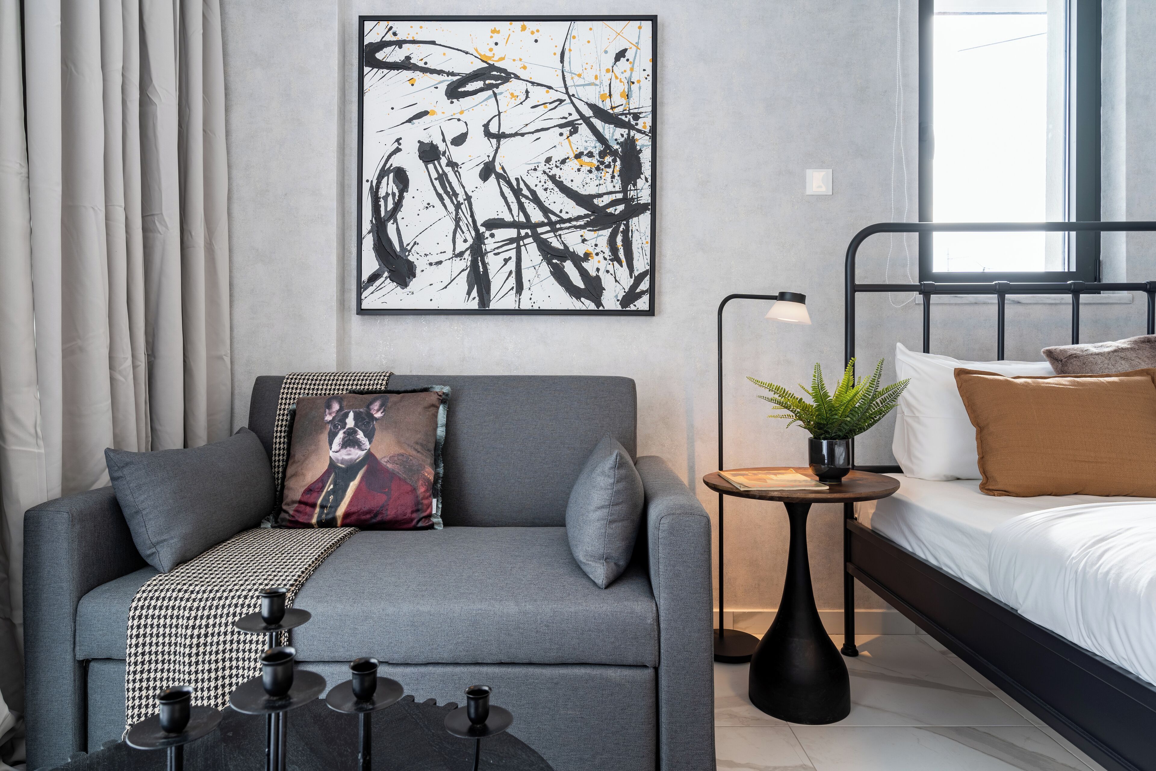A modern bedroom with a grey sofa, a black metal bed frame, a bedside table with a plant, and a framed abstract painting on the wall.