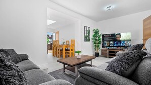 Una sala moderna con sofás grises, una mesa de centro de madera y un televisor que muestra un partido. Se ve un comedor con sillas de madera y una mesa al fondo.