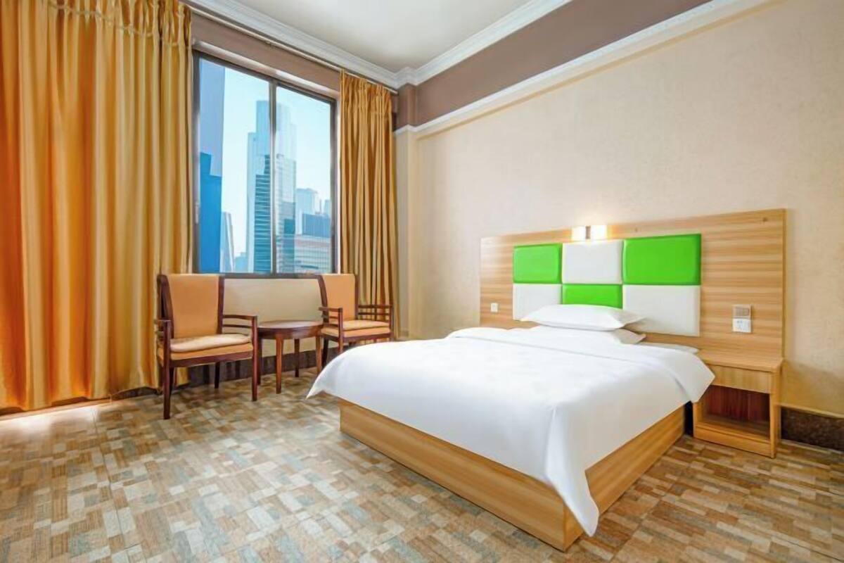 A hotel room with a bed, two chairs, a small table, a city view, and a window with curtains.