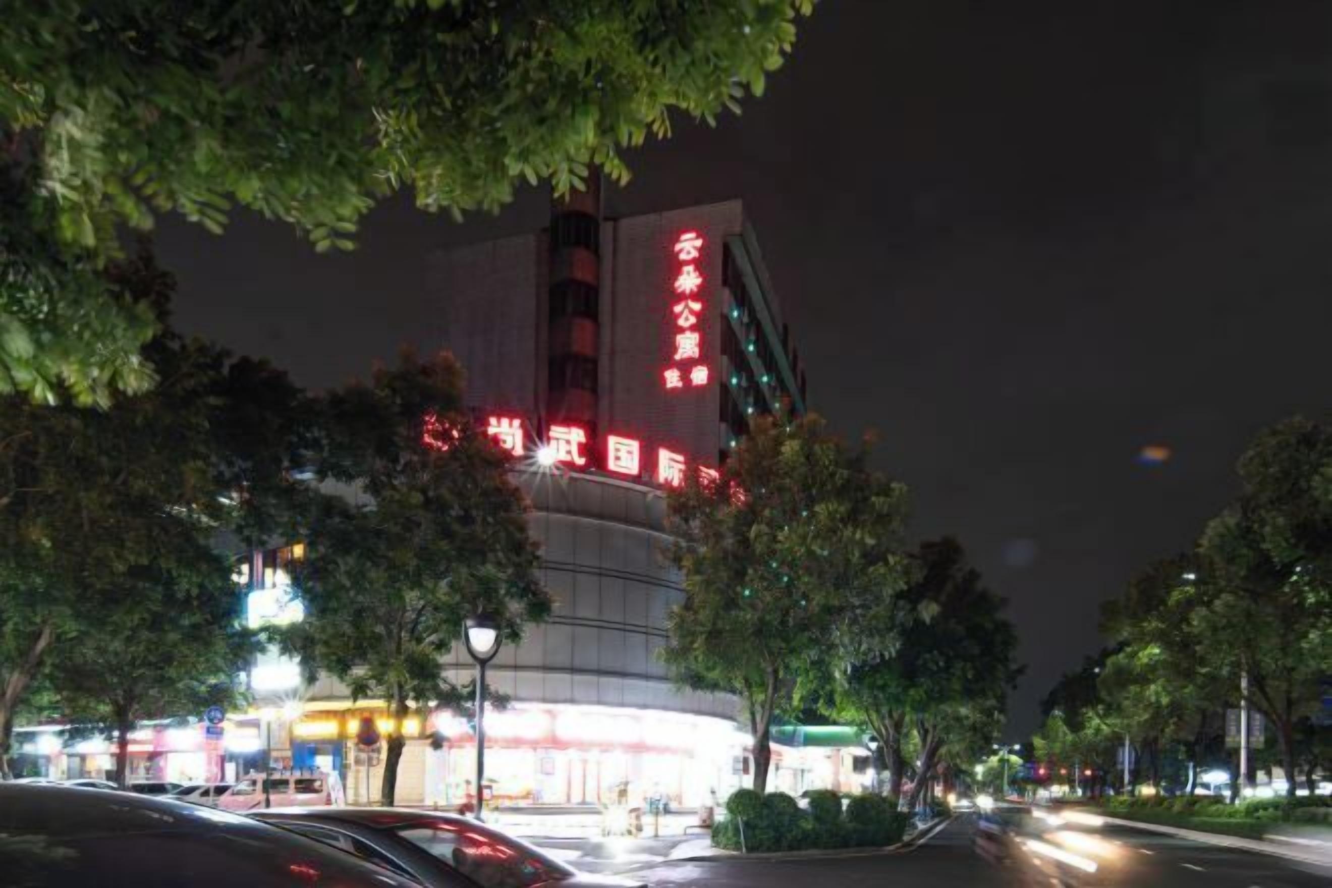 A building with illuminated signage at night, surrounded by trees and a street with moving vehicles.
