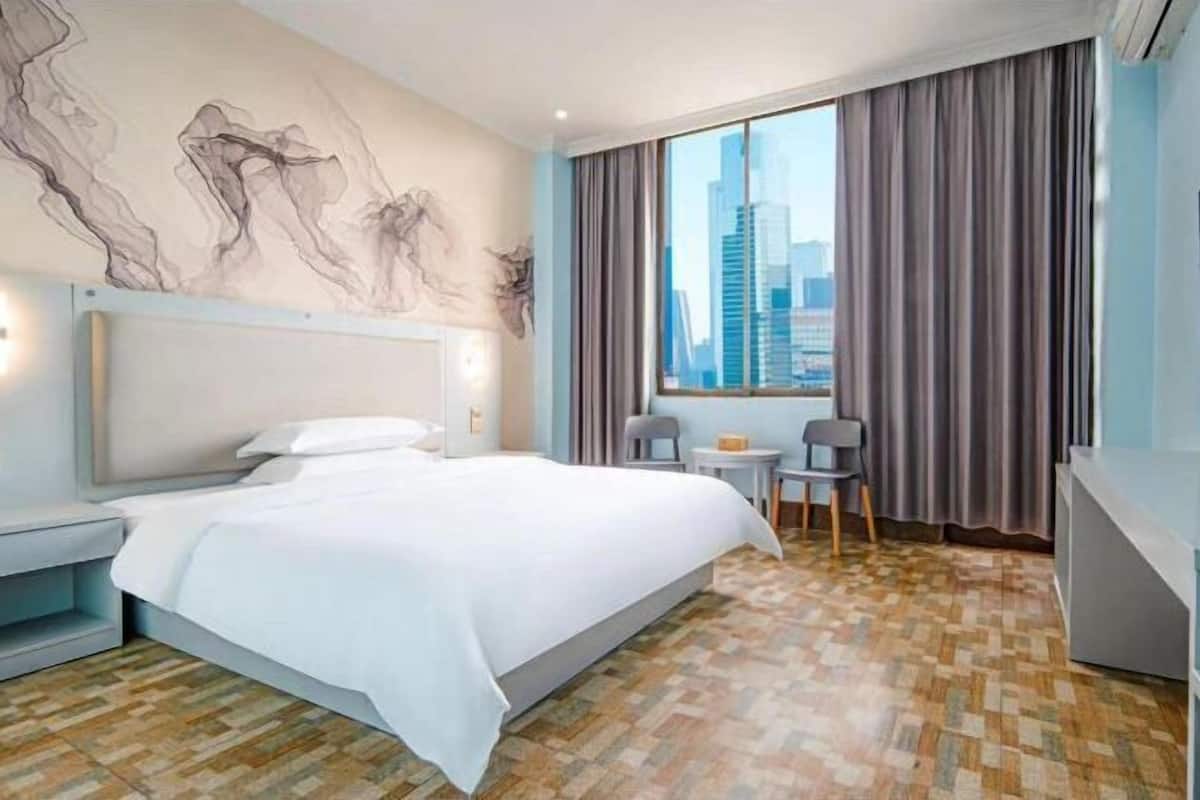 A hotel room with a bed, a desk, a chair, a view of a cityscape, and a wall with abstract art.