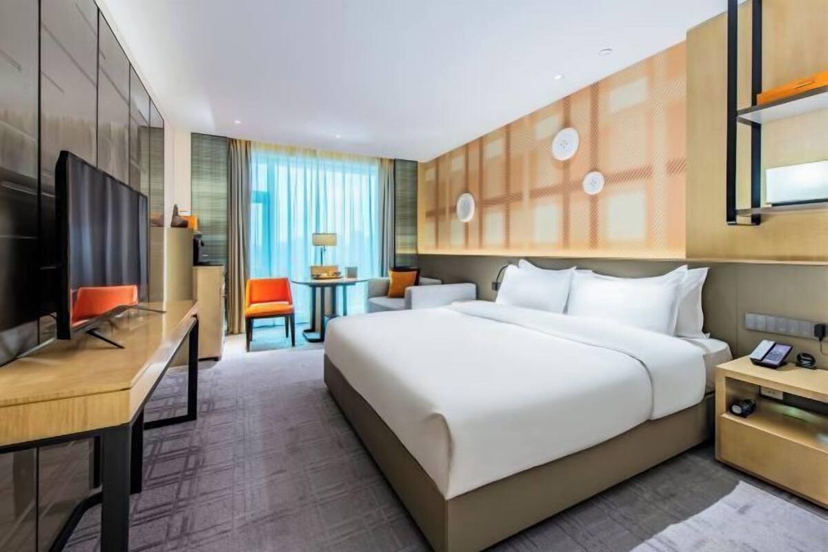 A hotel room with a large bed, a desk, a chair, a TV, and a small table with a lamp.