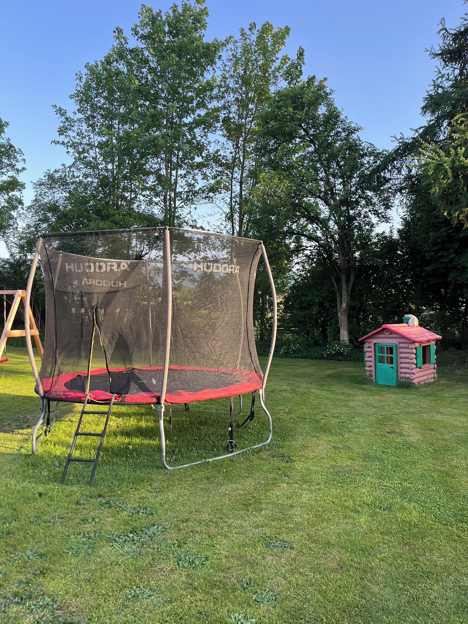 A trampoline with a safety net, a small pink playhouse, and a wooden swing set in a grassy area with trees in the background.