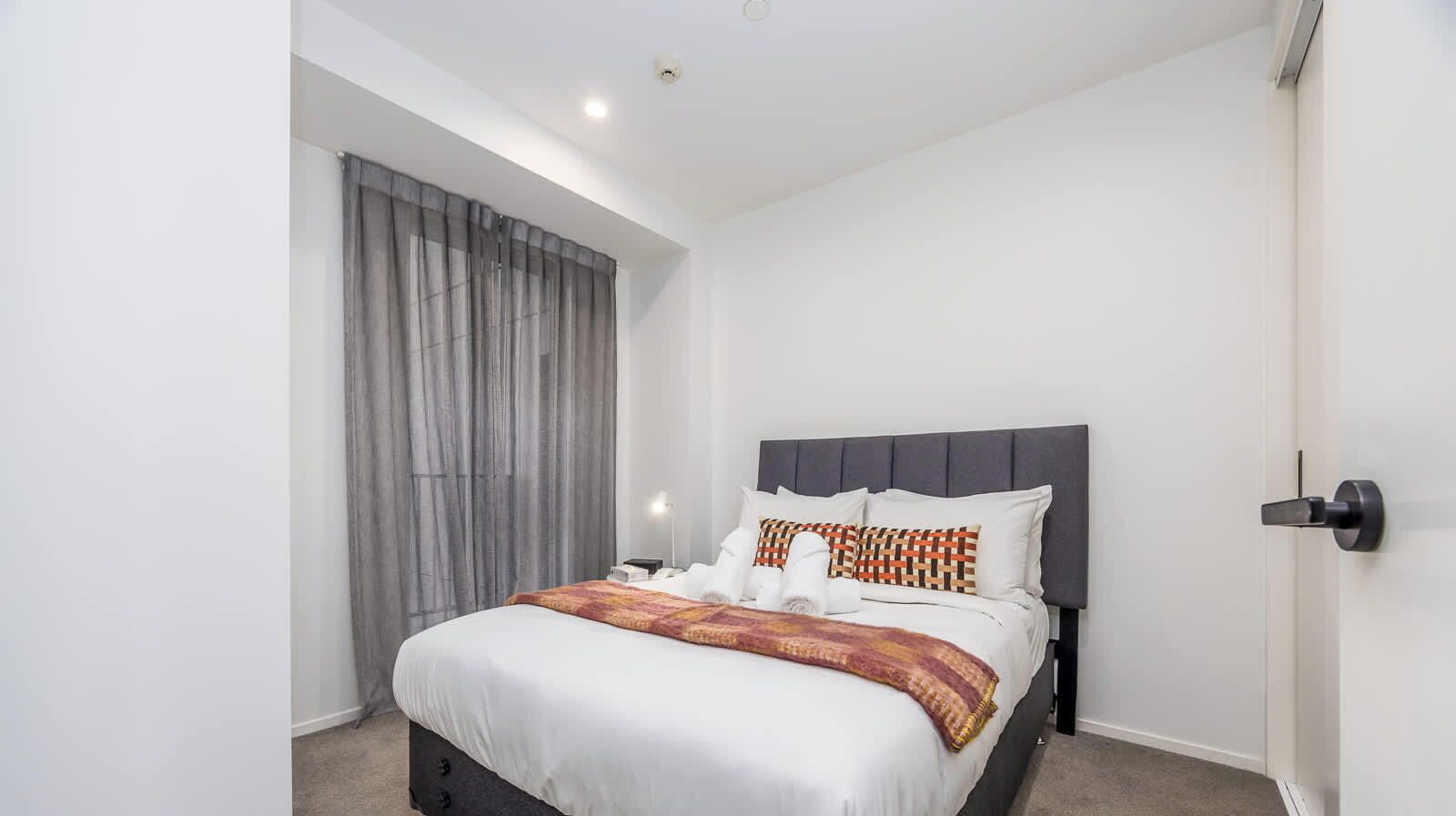 Una habitación de hotel moderna con una cama grande, cortinas grises, una mesita de noche con lámpara y un cabecero oscuro.