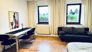 Ein modernes Schlafzimmer mit Holzboden, einem Bett, einem Schreibtisch mit zwei Stühlen, einem schwarzen Sofa und einem Fenster mit Vorhängen.
