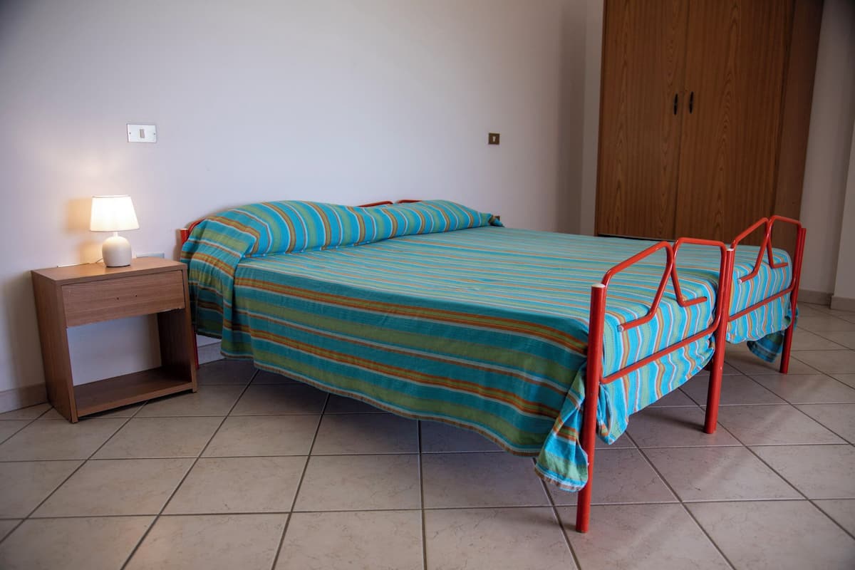 Un lit simple avec une couvre-lit à rayures, une table de chevet avec une lampe et une armoire en bois dans une pièce dont le sol est carrelé.