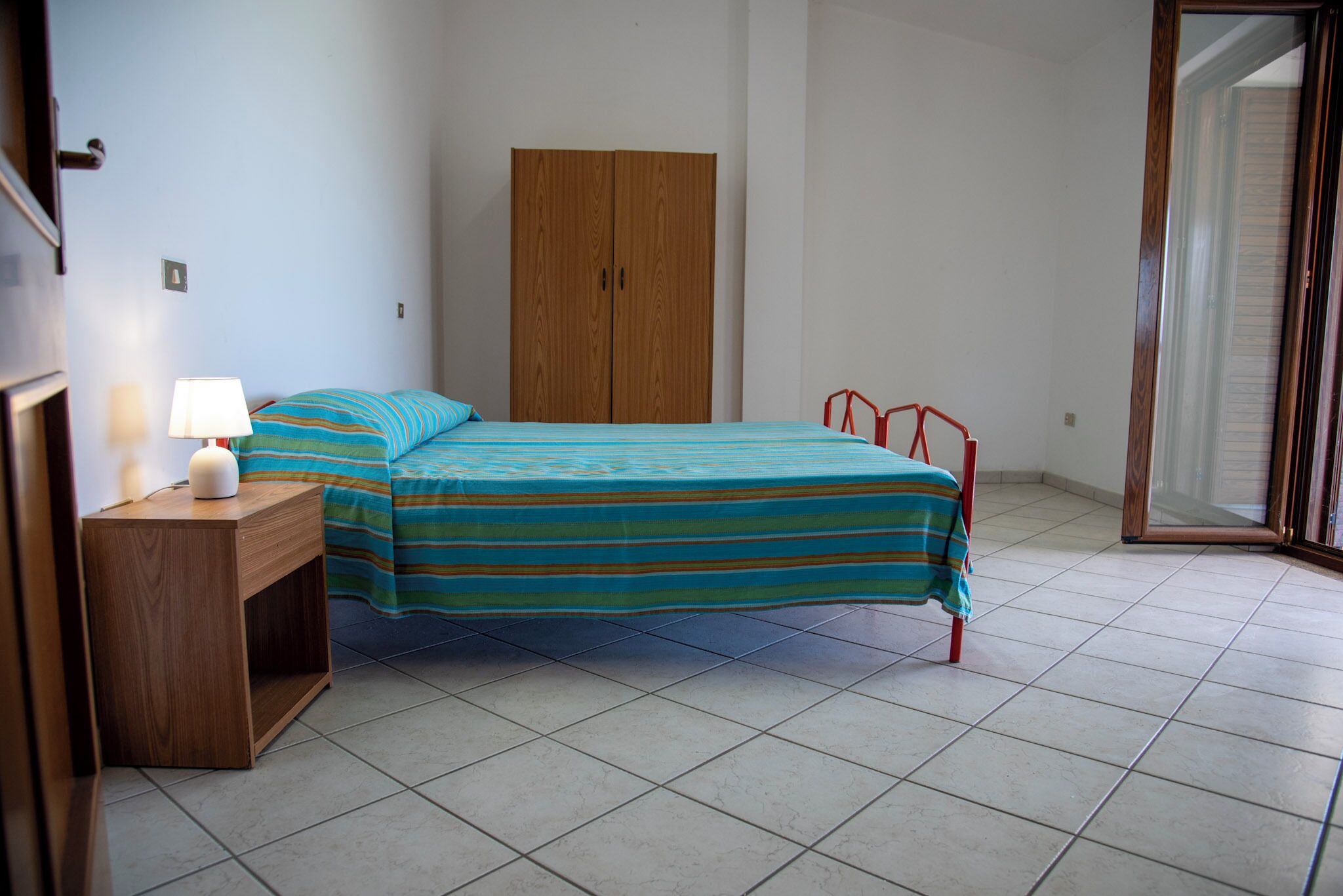 Une chambre à coucher avec un lit, une armoire en bois, une table de chevet avec une lampe et une fenêtre avec des stores.