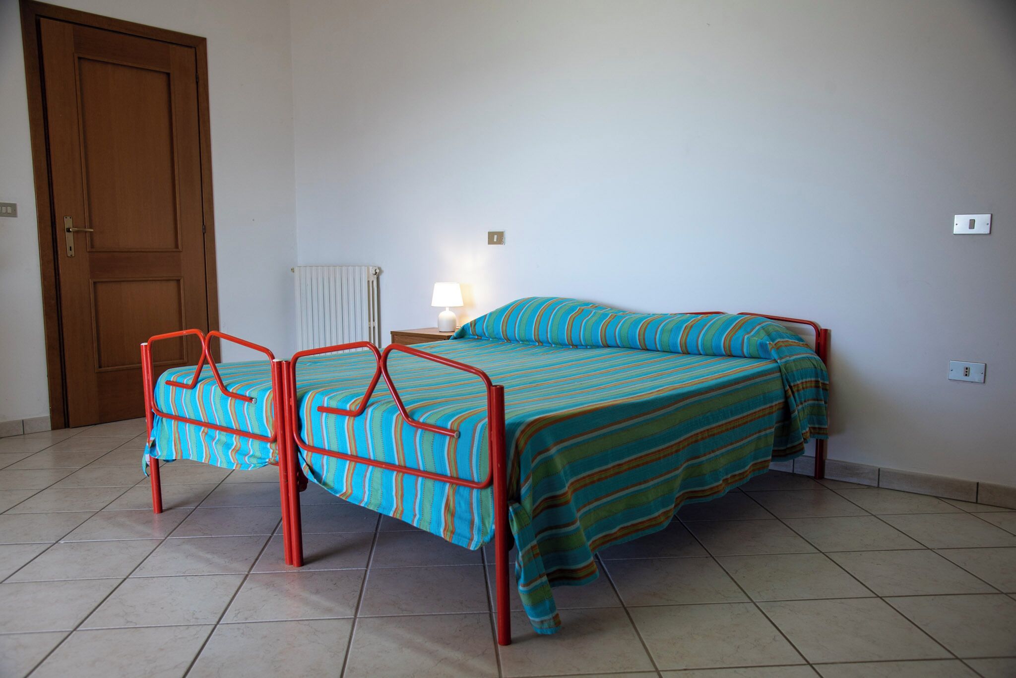 A bedroom with two single beds, a nightstand with a lamp, a radiator, and a wooden door.