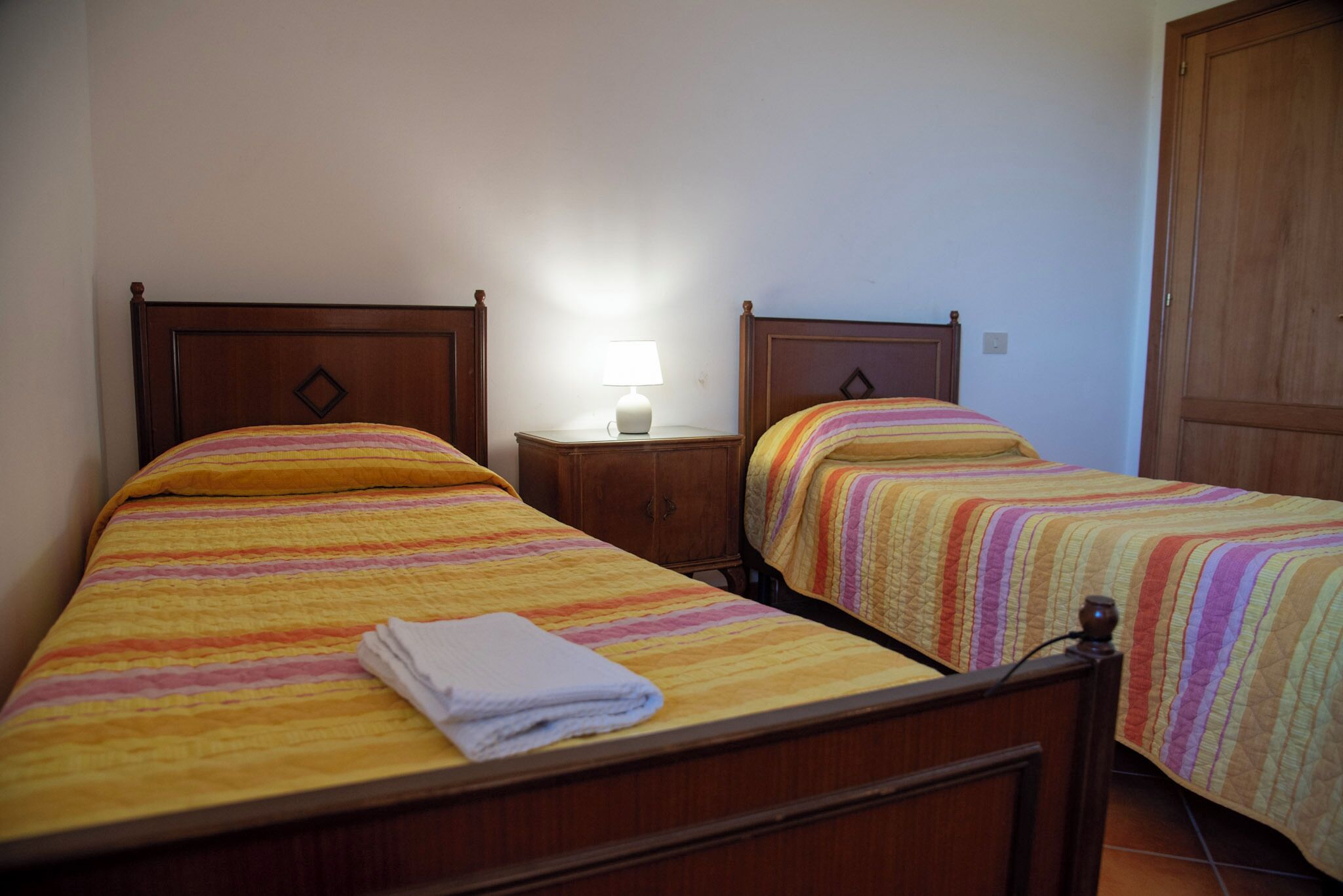 Une chambre d’hôtel avec deux lits simples, chacun doté d’une tête de lit en bois et d’une couvre-lit rayée aux couleurs vives. Une petite table de chevet avec une lampe se trouve entre les deux lits.