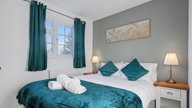 A neatly made bed with a teal blanket, two bedside lamps, and a window with curtains. - CircleWay Homes - Colwick (Colwick)