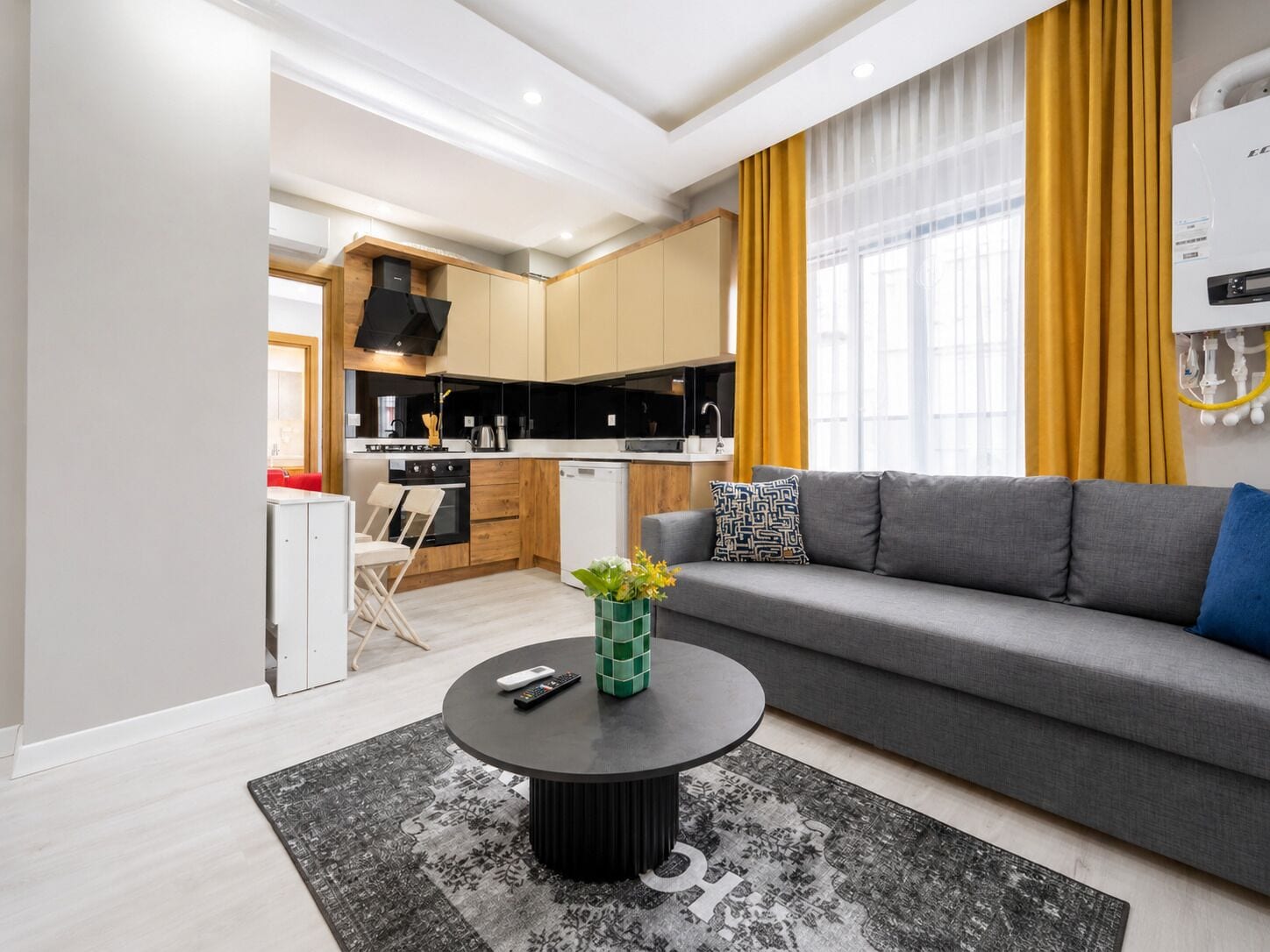 A modern living room with a grey sofa, a round coffee table, and a kitchen area with wooden cabinets and a black range hood.