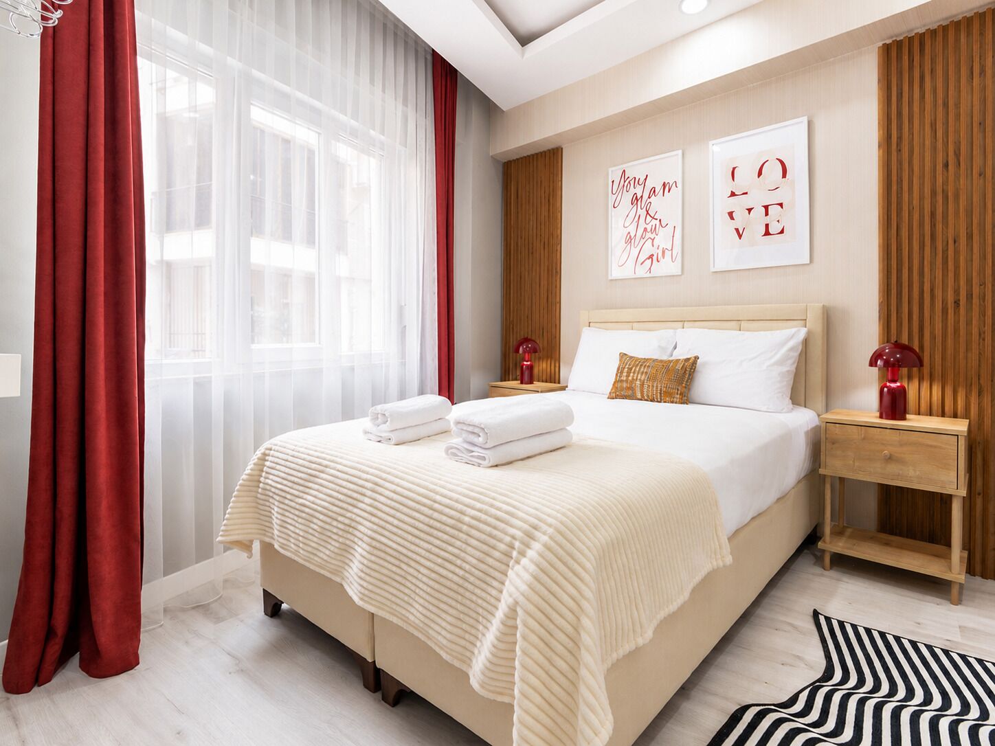 A hotel room with a bed, two bedside tables, a nightstand, a window with sheer curtains, and artwork on the wall.