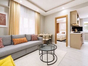 Una habitación de hotel moderna con un sofá gris, una mesa de centro de vidrio, una zona de comedor con una mesa pequeña y sillas, una kitchenette y una cama con edredón blanco.