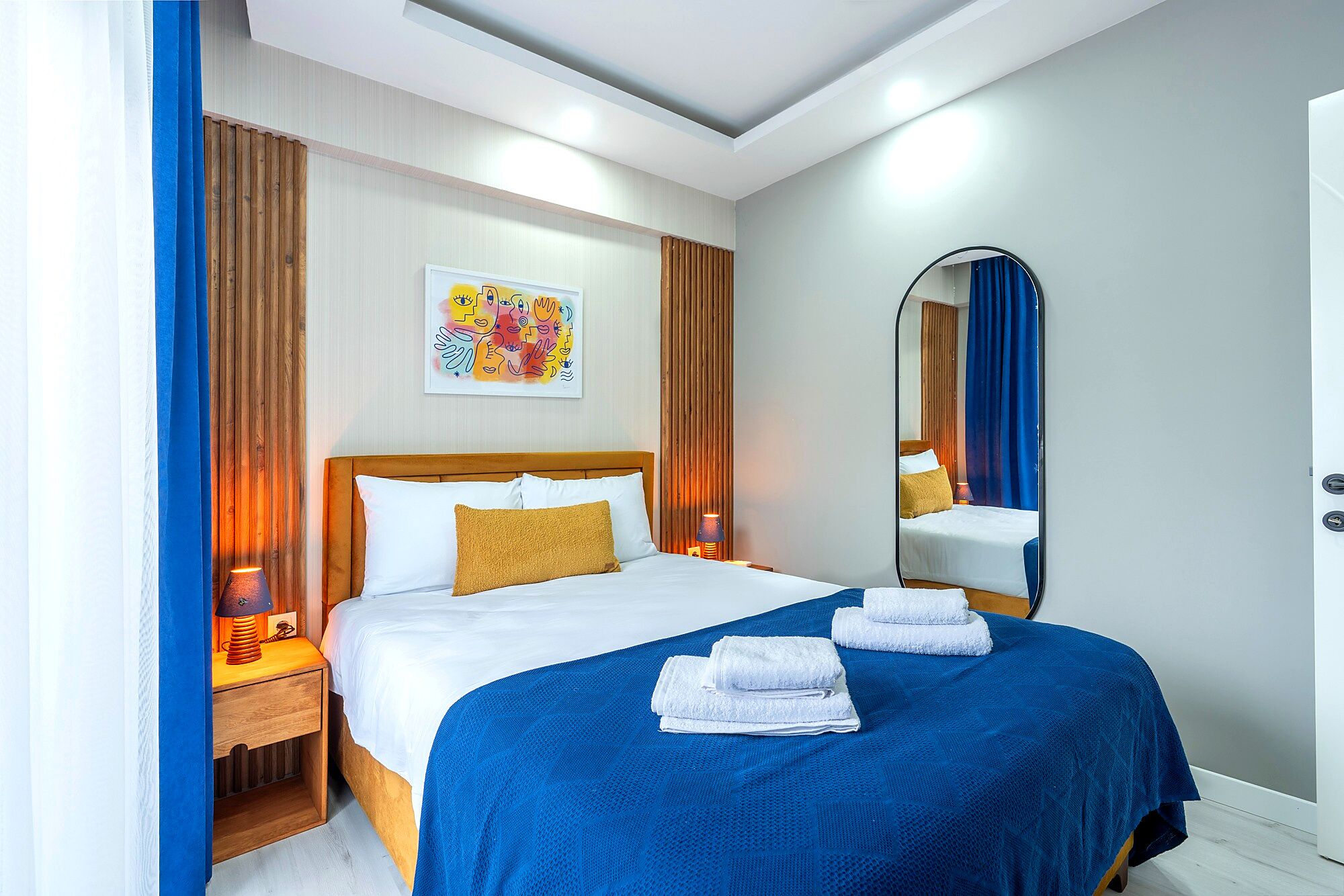 A hotel room with a bed, a nightstand, a lamp, a mirror, and a blue curtain.