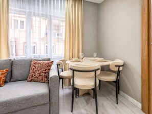 Una sala moderna con un sofá gris, cojines estampados, una mesa redonda de comedor con sillas y vistas a edificios a través de amplios ventanales.