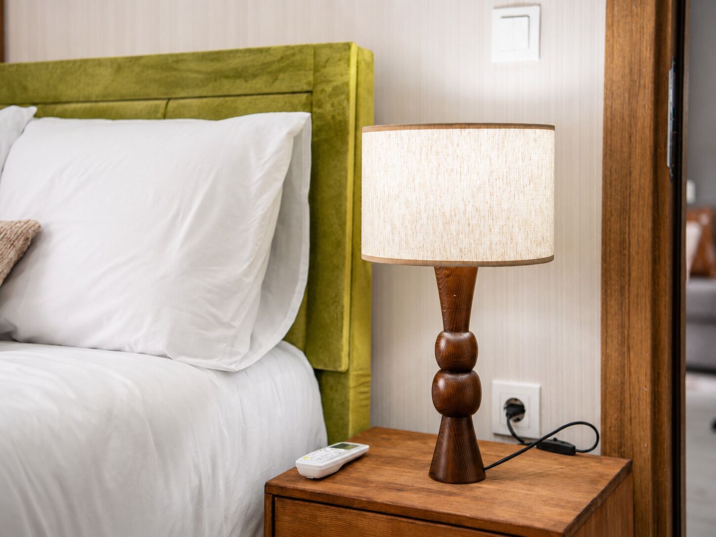 A neatly made bed with white linens and a green headboard, a wooden bedside table with a lamp, and a remote control.