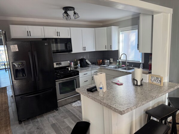 A modern kitchen with white cabinets, stainless steel appliances, a granite countertop, and a small island with stools. - River Trail Retreat by the Bay (Bay City)