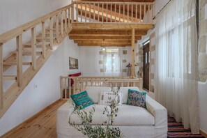 A wooden staircase leading to an upper level, a white sofa with patterned cushions, and a window with sheer curtains. - Kalimera Loft I – Village Experience ,a cozy retreat (Argiroupoli)