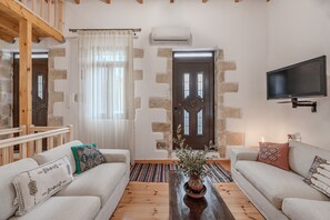 A living room with a sofa, a coffee table, a TV, and a door with a stone wall. - Kalimera Loft I – Village Experience ,a cozy retreat (Argiroupoli)
