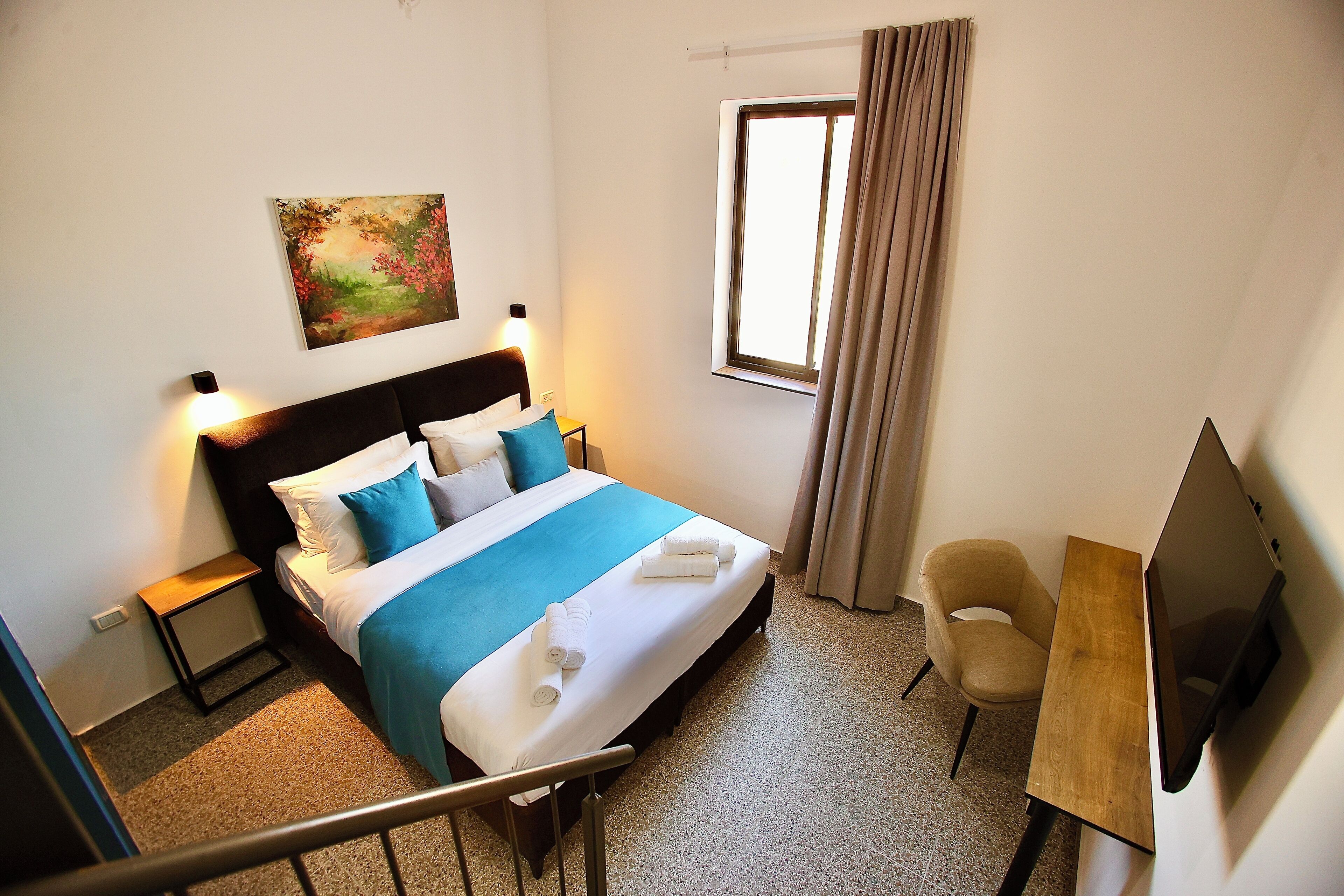 A hotel room with a bed, a desk, a chair, a TV, a painting on the wall, and a window with curtains.