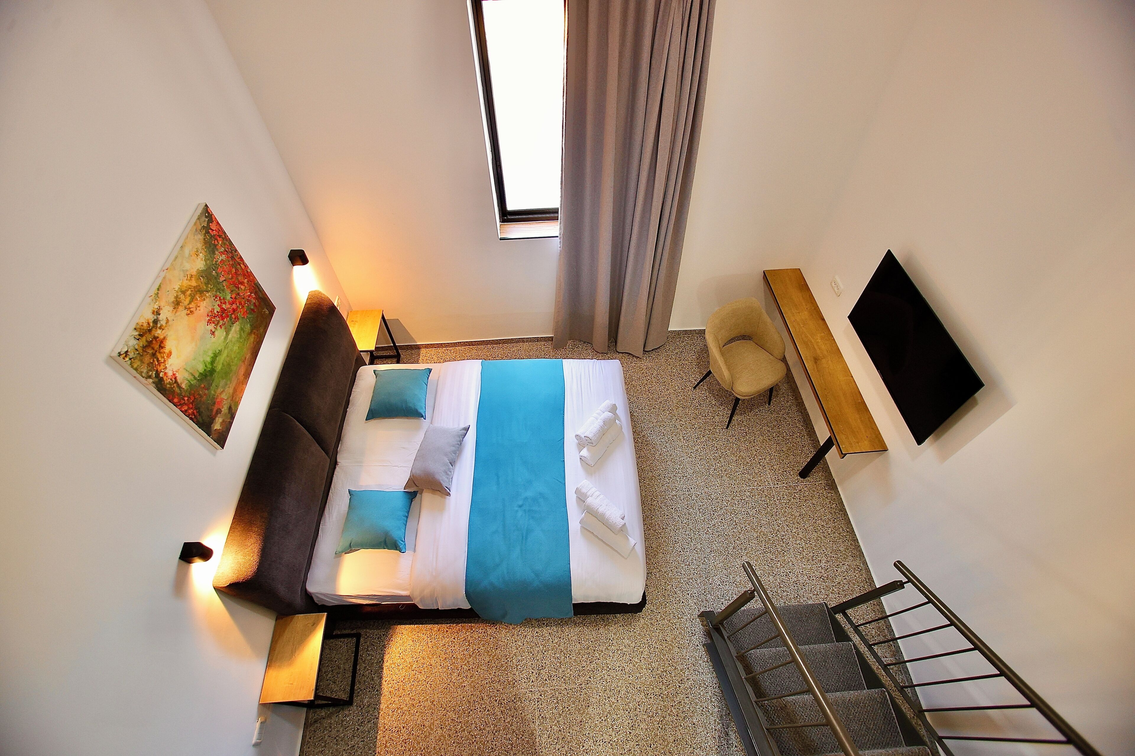 A hotel room with a bed, a painting, a desk, a chair, a TV, and a window with curtains.