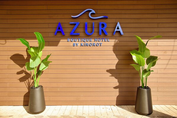 Un letrero que dice 'AZURA Boutique Hotel by Kinorot' con dos plantas en macetas al frente.