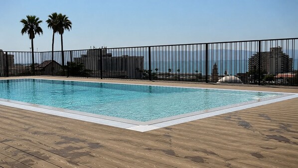 Una piscina rectangular con un muelle de madera alrededor, una cerca metálica y palmeras al fondo.